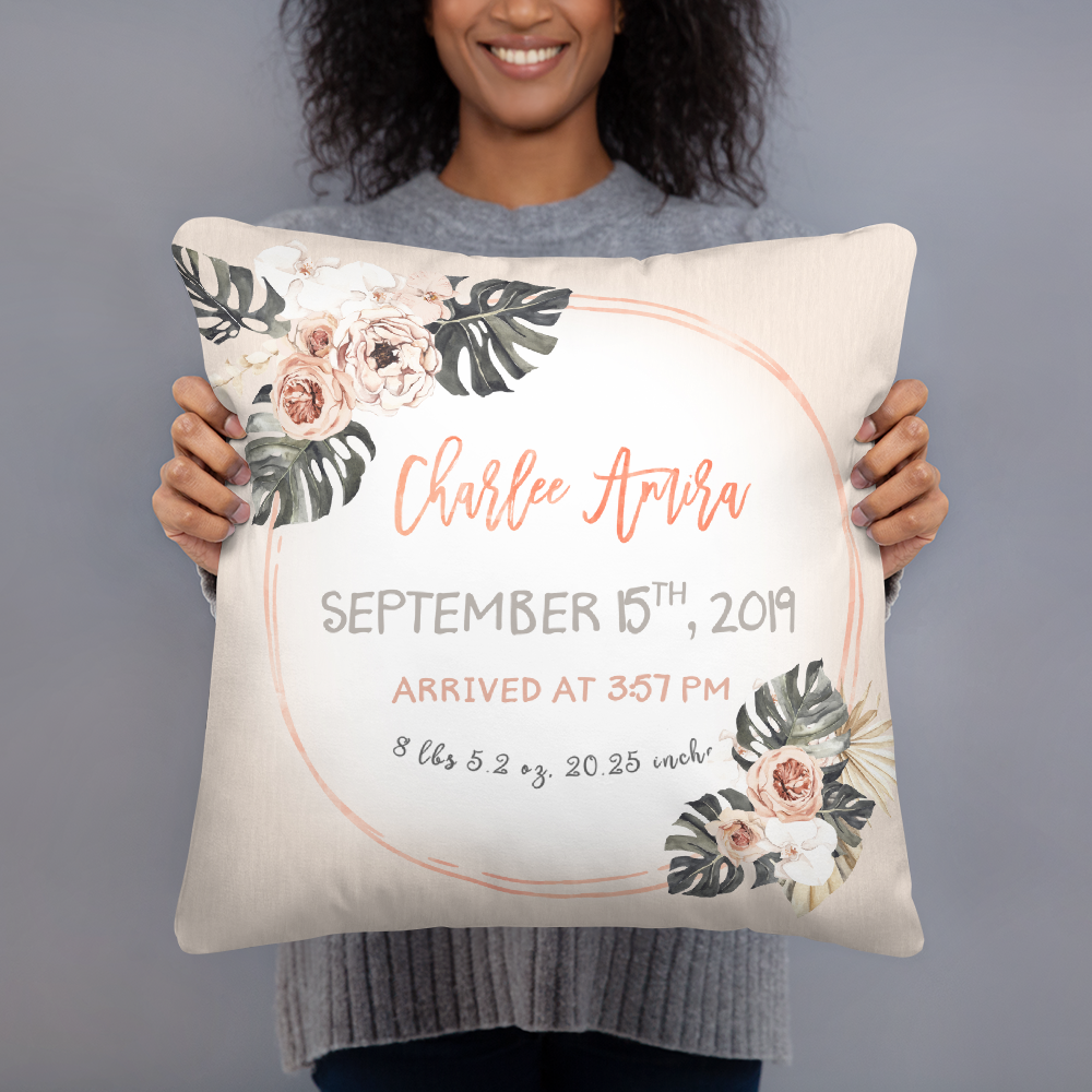 The person holding a modern tropics themed birth announcement pillow that says the baby's name, date, time, weight, and length. It is orange and beige with flowers and leaves.